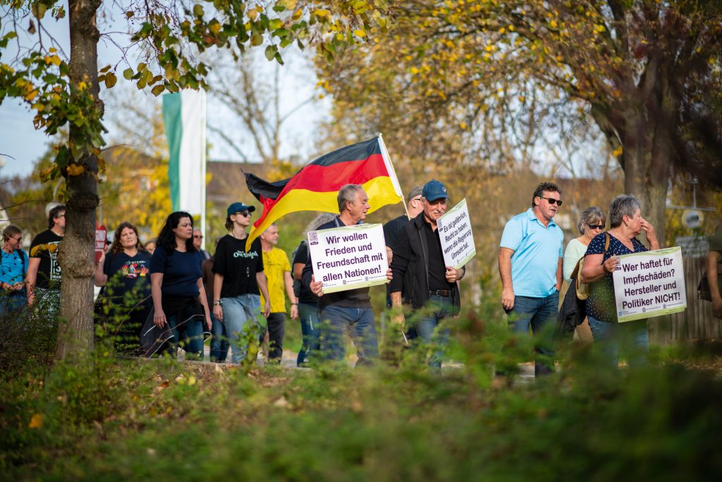 Demonstrierende mit Deutschlandflagge. Auf ihren Schildern steht "Wir wollen Frieden und Freundschaft mit allen Nationen!" oder "Wer haftet für Impfschäden? Hersteller und Politiker NICHT!"